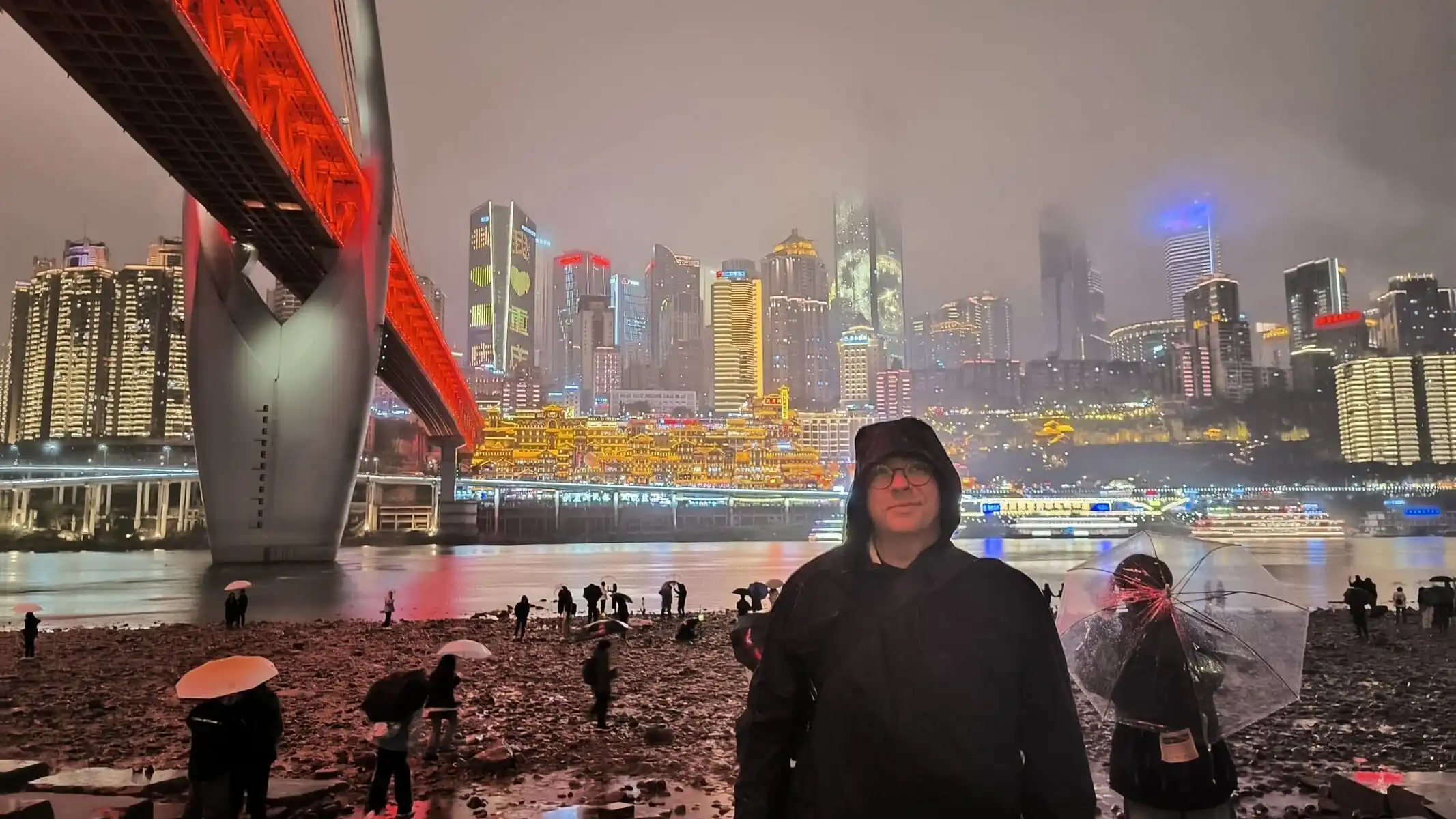 Qiansimen Jialing River Bridge, Hongya Cave, chongqing, chinatrip, Night city skyline at night with a bright red-lit bridge, misty skyline, and people on a rocky shore holding umbrellas in the rain.