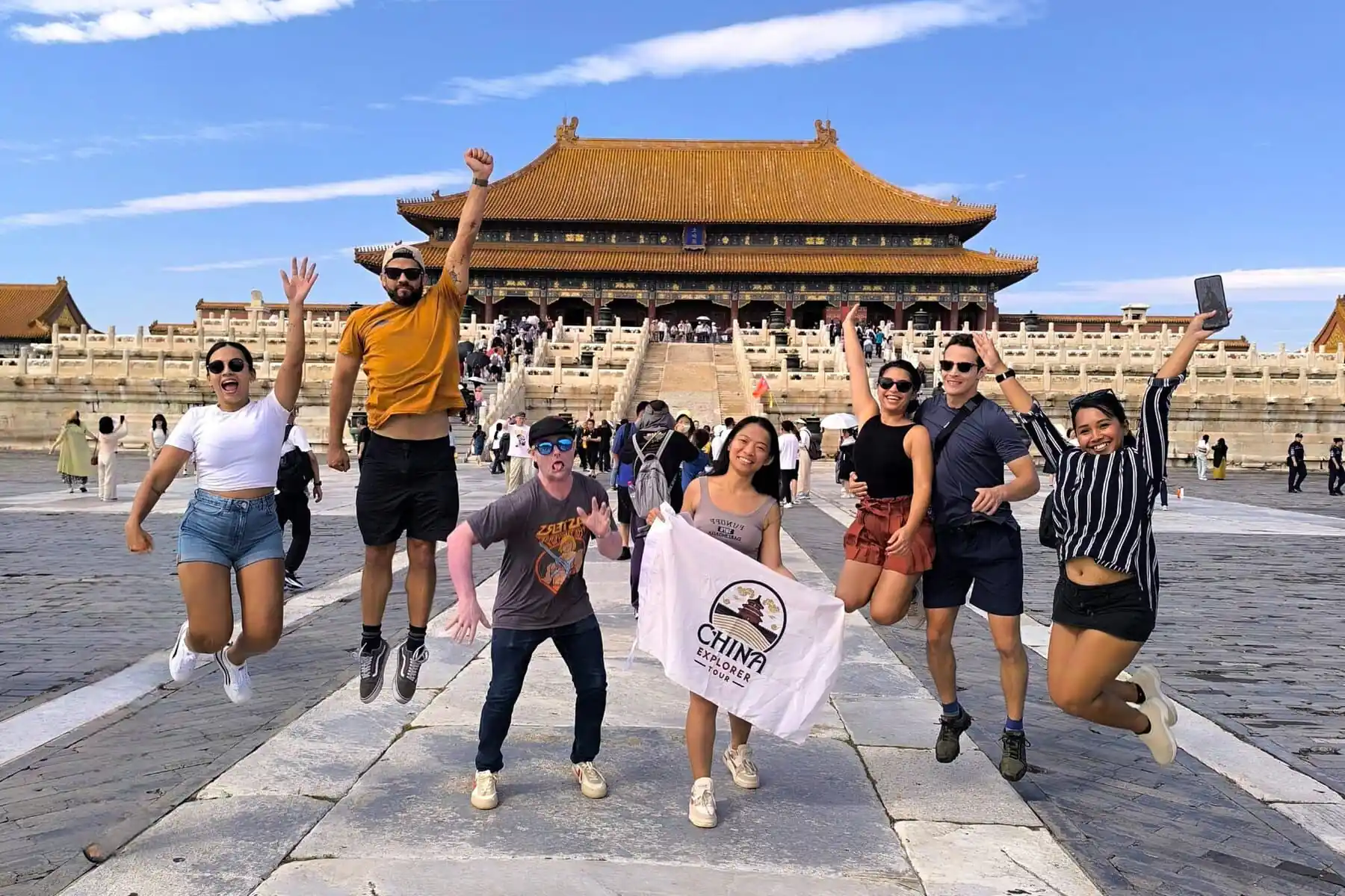 the forbidden city, beijing, chinatrip, Group of seven tourists mid-jump in front of a yellow-roofed imperial hall; one holds a China Explorer Tour banner.