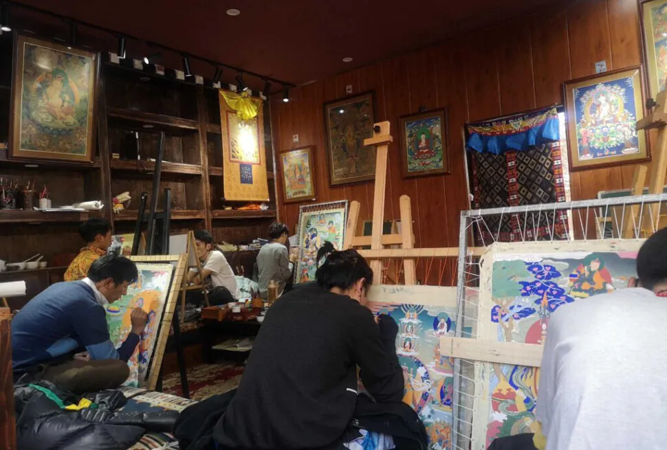 tangka, tibet, chinatrip, Art studio with several people painting on large colorful canvases, surrounded by wooden walls, framed artworks, and easels; artists are seated on the floor or on cushions working intently.