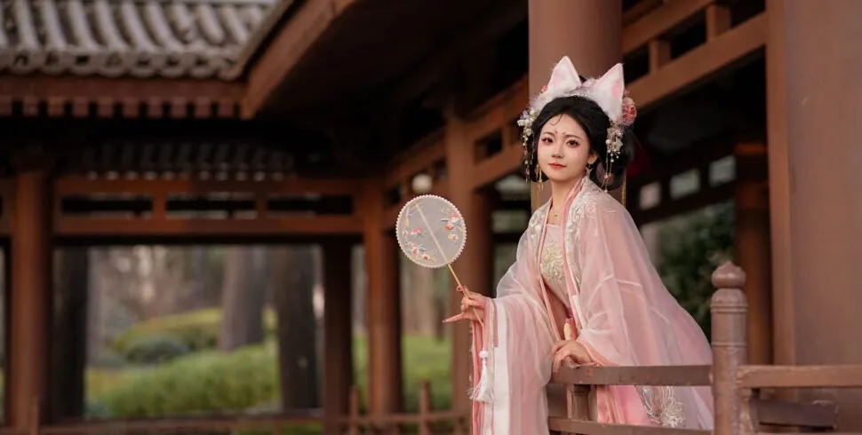 hanfu photoshoot, chinatrip, Young woman in a pale pink traditional dress with floral embroidery, standing on a wooden veranda and holding a round decorative fan.