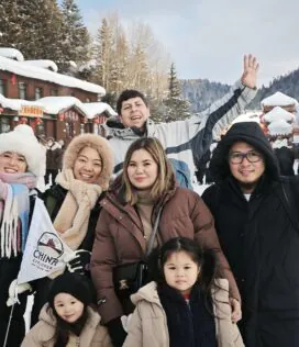 Harbin Trip, heilongjiang, china tour, Group of friends smiling and waving in a snowy village street with wooden shops and red lanterns.
