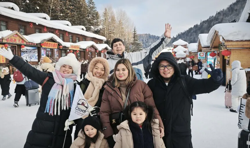 Harbin Trip, heilongjiang, china tour, Group of friends smiling and waving in a snowy village street with wooden shops and red lanterns.