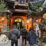 Arhat temple, Chongqing, china tour, People wearing winter coats walk toward a traditional wooden temple gate with ornate carvings and a blue sign, warm orange lantern light inside.