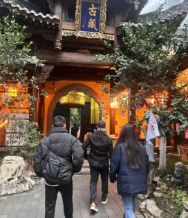 Arhat temple, Chongqing, china tour, People wearing winter coats walk toward a traditional wooden temple gate with ornate carvings and a blue sign, warm orange lantern light inside.