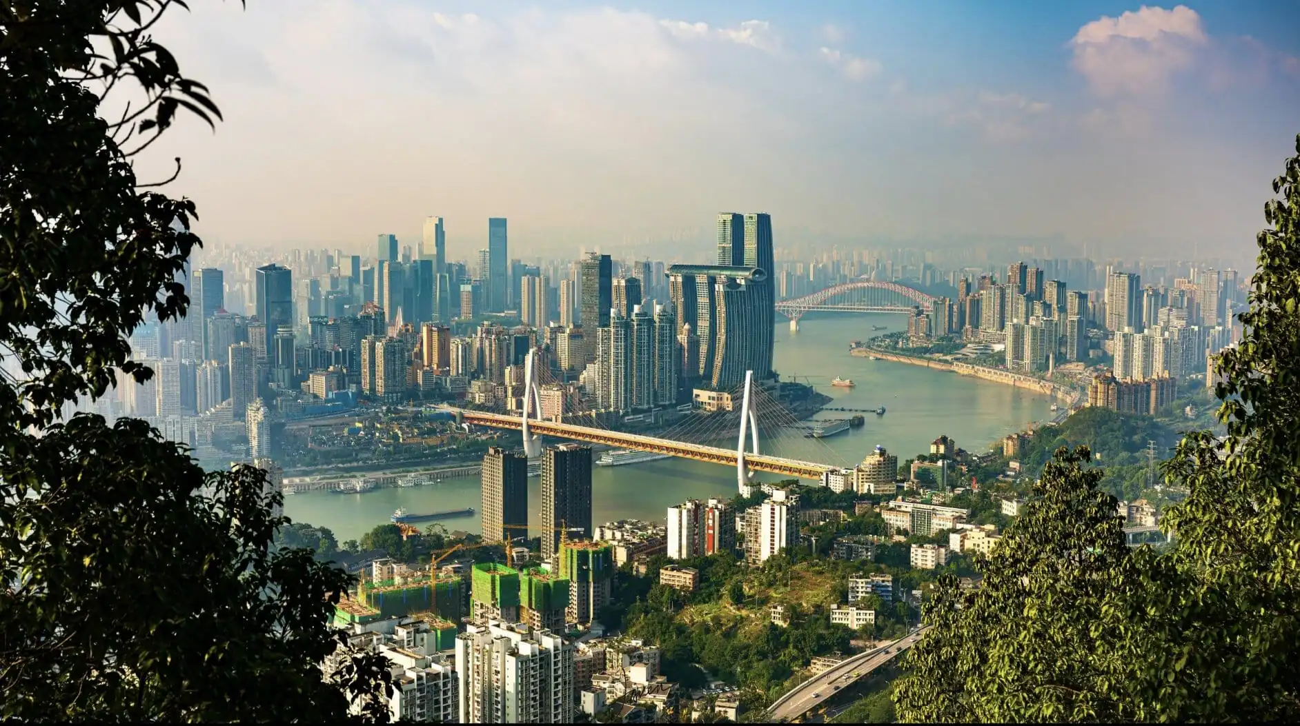 Panoramic cityscape with high-rise buildings along a winding river, framed by green hills and trees in the foreground. Two bridges span the river among dense skyscrapers.",