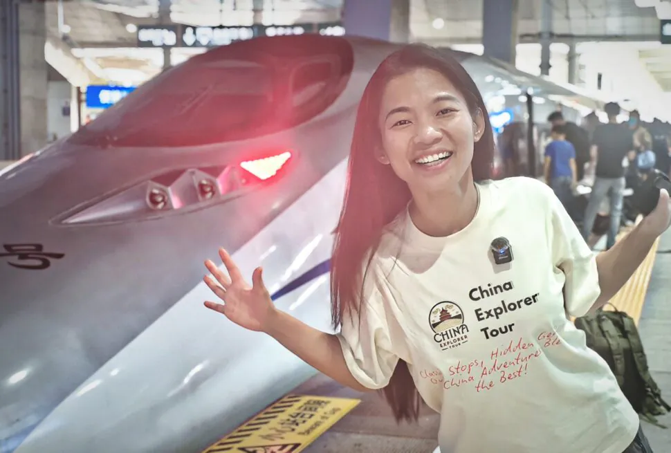 china fast train, chinatrip, Woman smiles and poses beside a fast train at a station, wearing a white shirt with 'China Explorer Tour' text.