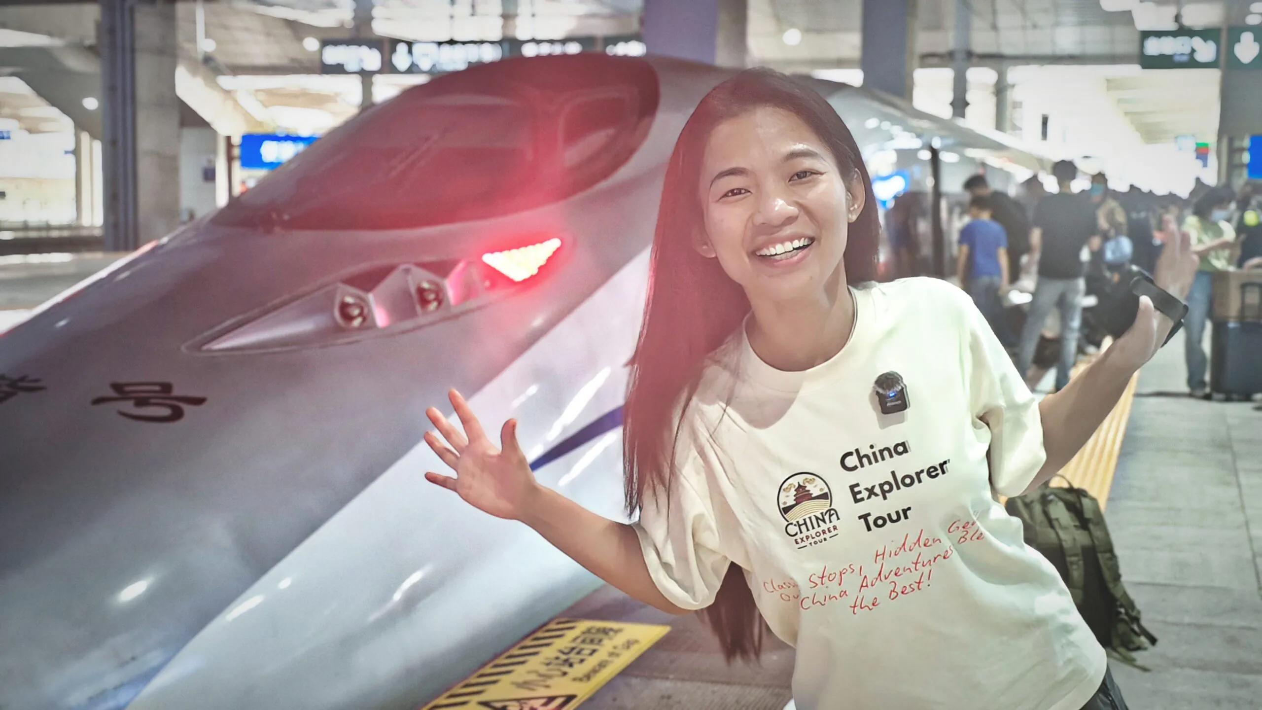 china fast train, chinatrip, Woman smiles and poses beside a fast train at a station, wearing a white shirt with 'China Explorer Tour' text.