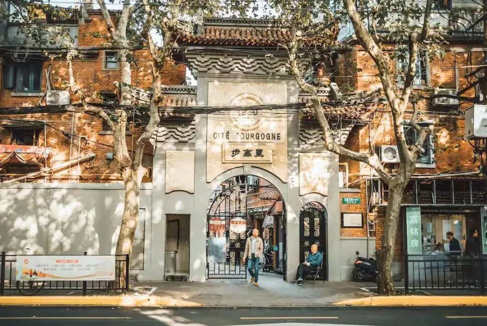 CITE BOURGOGNE, Shanghai, chinatrip, Entrance arch labeled 'Cité Bourgogne' in a brick-walled street, with trees and pedestrians nearby on a sunny day.