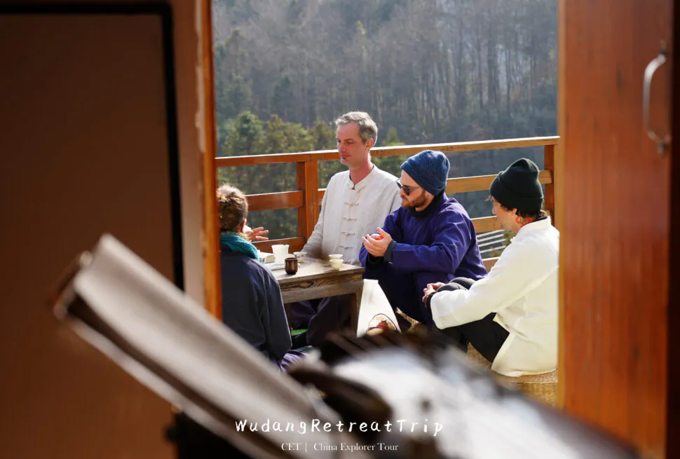 wudang, taichi, chinatrip, Group of people sitting around a wooden table on a sunlit veranda, drinking tea with a forest backdrop behind them.