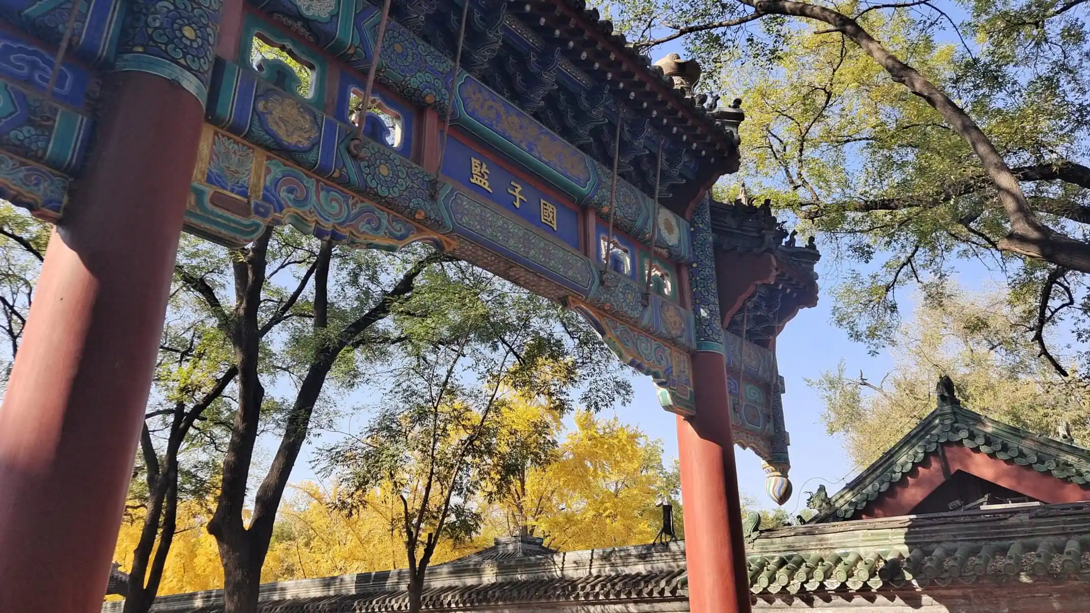 imperial college, beijing, chinatrip, Colorful Chinese temple archway with red columns and intricate blue-green carvings against a blue sky and autumn trees with yellow leaves.