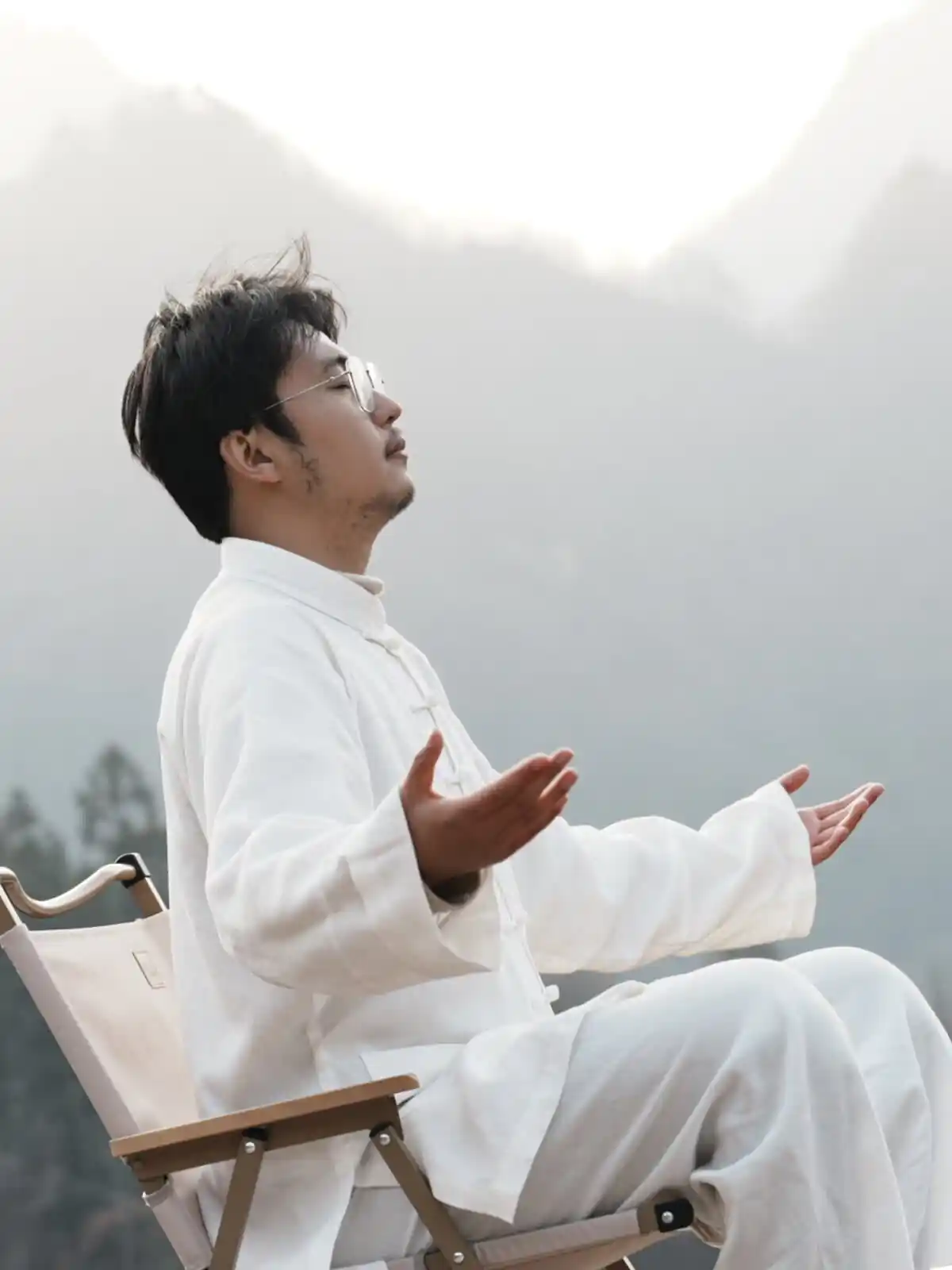 Man in white traditional clothing sits in a chair with arms open and eyes closed, meditating outdoors with misty mountains in the background.