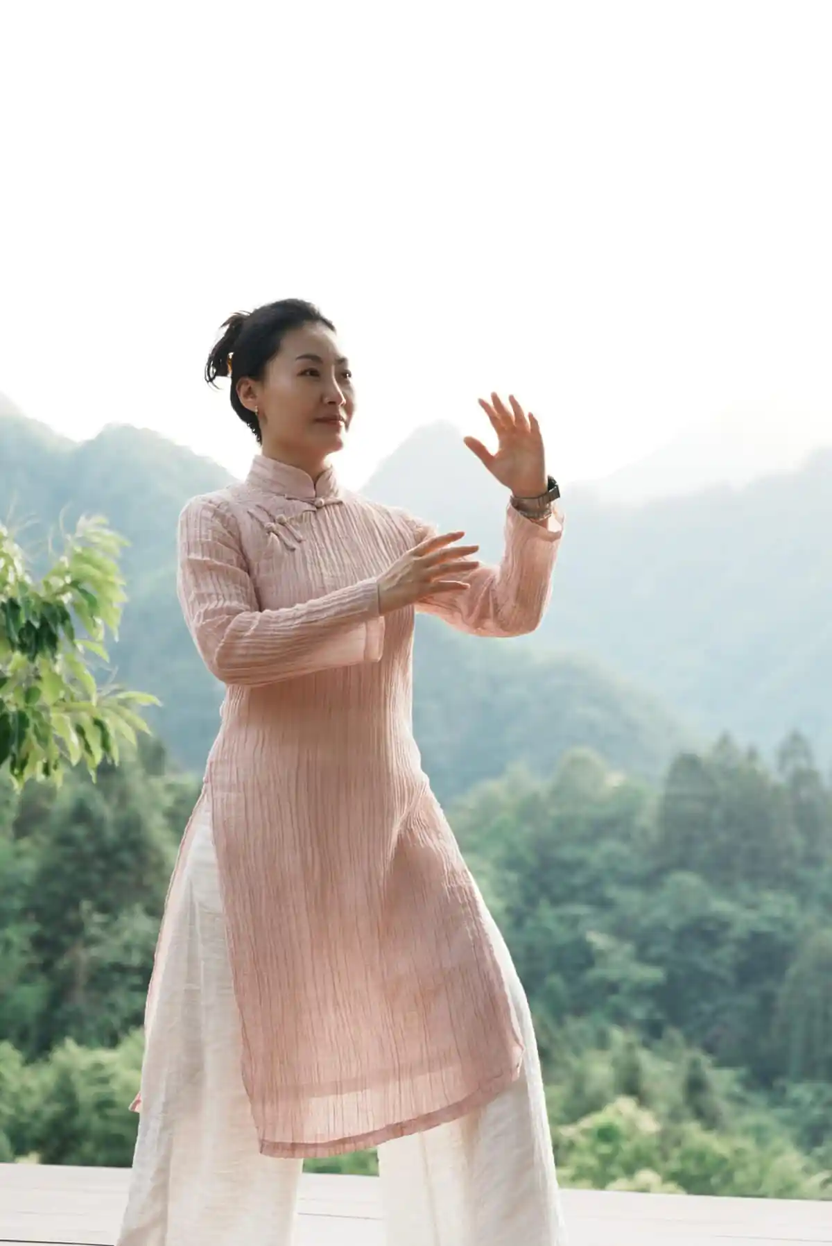 Malena, China Explorer Tour, chinatrip, Woman in a pale pink traditional outfit performing a slow martial-arts pose outdoors with green hills in the background.