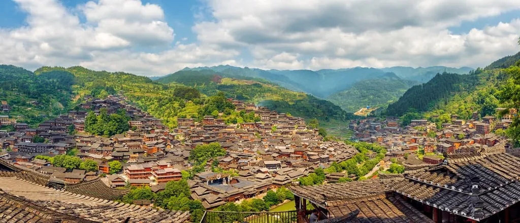 Panoramic view of a hillside village with densely packed wooden buildings and tiled roofs amid green mountains.