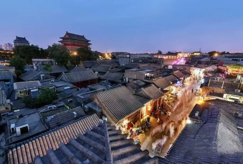 Beijing Hutong Tour,Chinatour Aerial view of a historic Chinese town at blue hour, with tiled roofs, a central pagoda, and a busy lantern-lit street below.