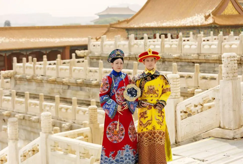 Two people in traditional Chinese imperial costumes pose on a stone bridge in a historic palace courtyard.