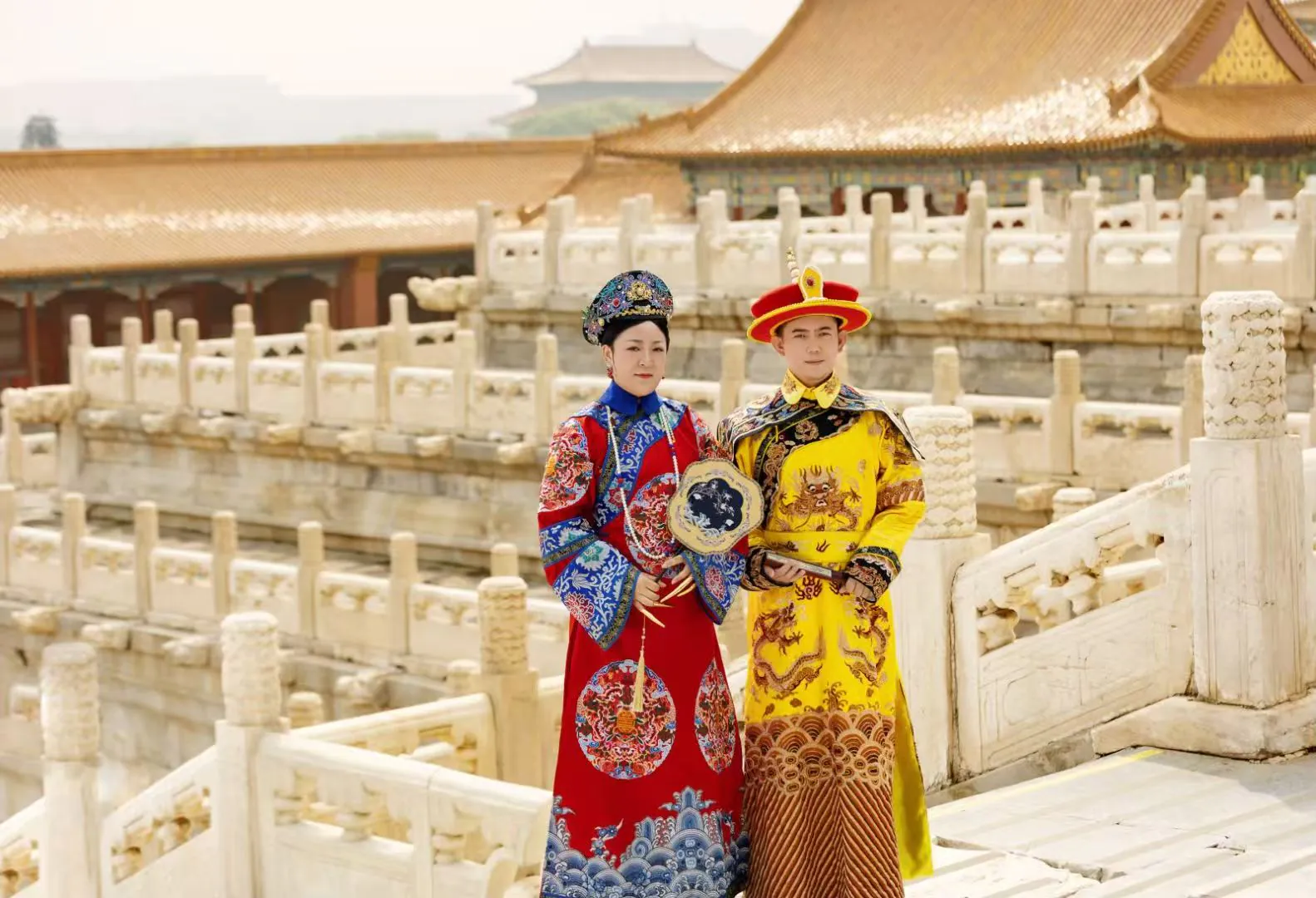Two people in traditional Chinese imperial costumes pose on a stone bridge in a historic palace courtyard.