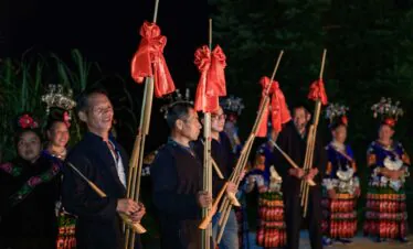 School trip in Guizhou, chinatrip, Group of people in traditional clothing holding tall bamboo poles with red fabric bows at a nighttime festival.