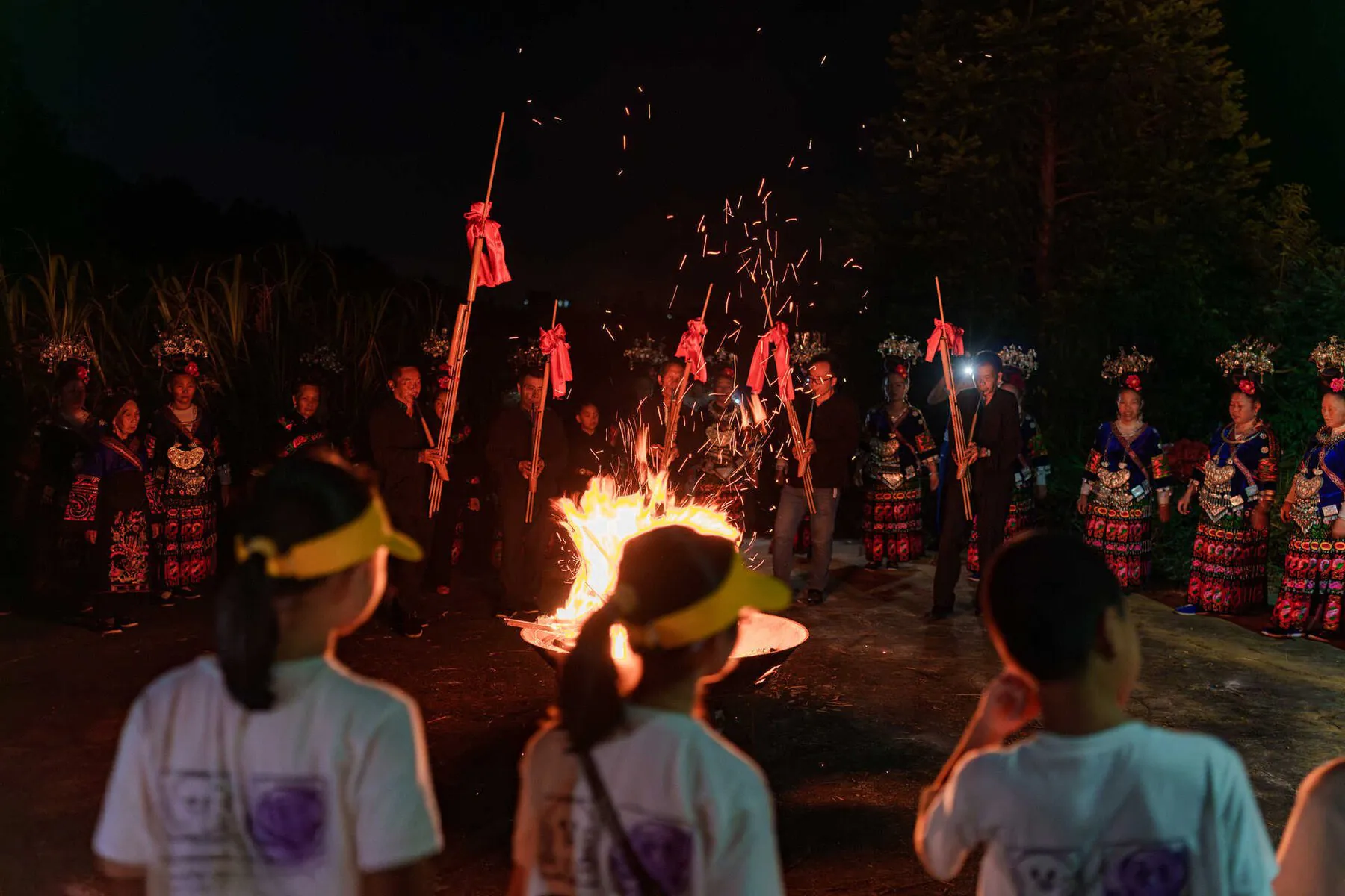 School trip in Guizhou, chinatrip, Group of people in colorful traditional costumes standing around a blazing bonfire at night, sparks flying from the flames.