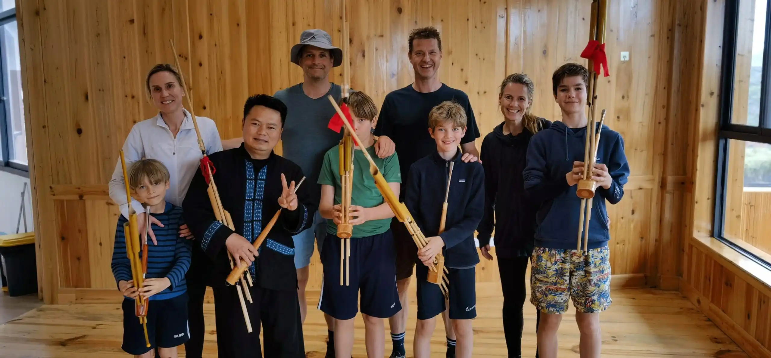 School trip in Guizhou, chinatrip, Group of nine people in a wood-paneled room holding bamboo wind instruments and smiling at the camera.