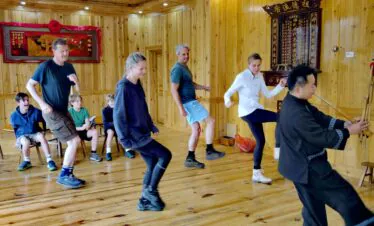 School trip in Guizhou, chinatrip, Group of people in a wooden studio dancing or practicing; a man at the far right plays a traditional erhu instrument while others watch or join in.
