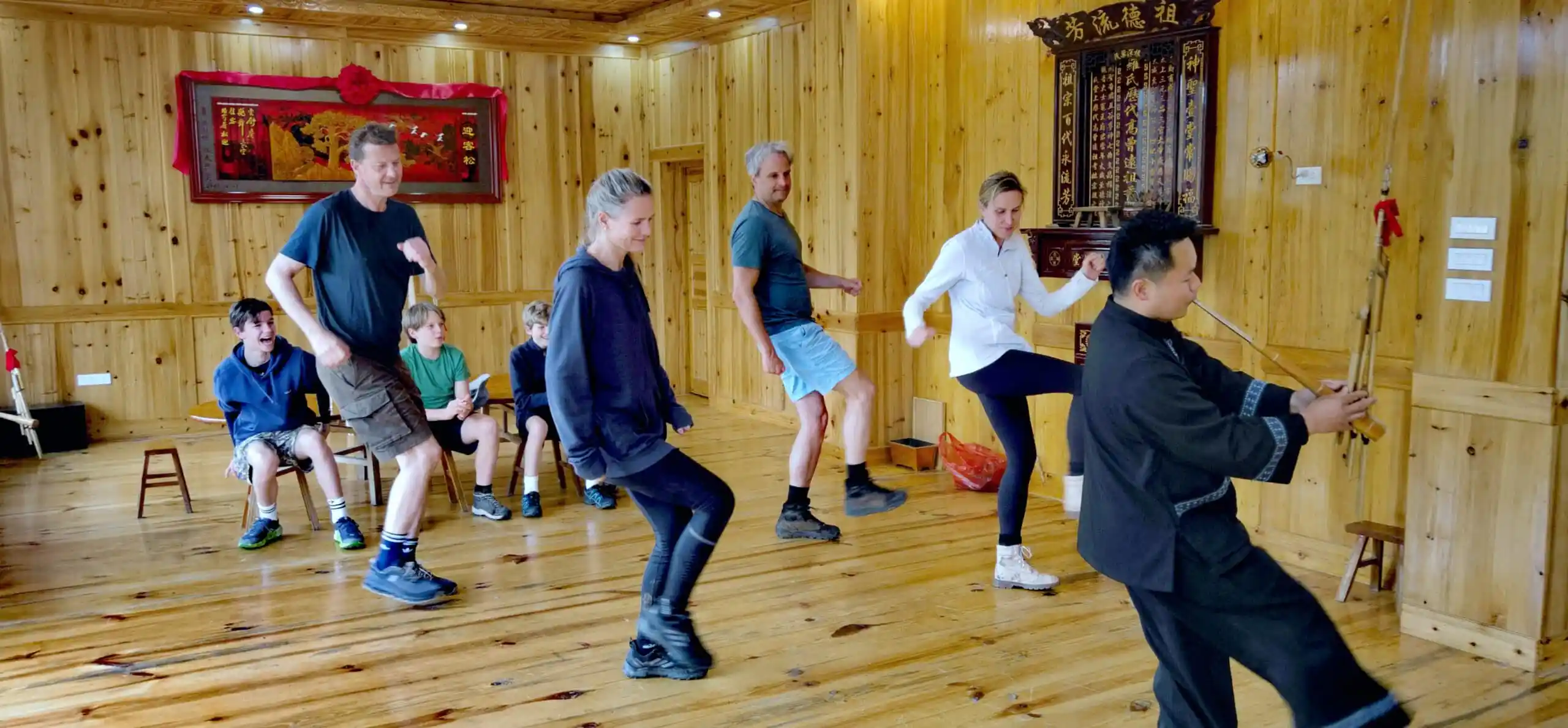 School trip in Guizhou, chinatrip, Group of people in a wooden studio dancing or practicing; a man at the far right plays a traditional erhu instrument while others watch or join in.