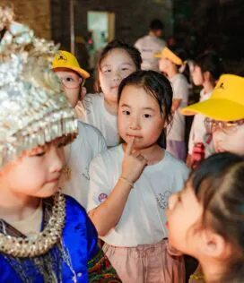 School trip in Guizhou, chinatrip, Children in a crowded festival, a girl in blue traditional dress with a beaded headdress among peers wearing yellow caps and white shirts.