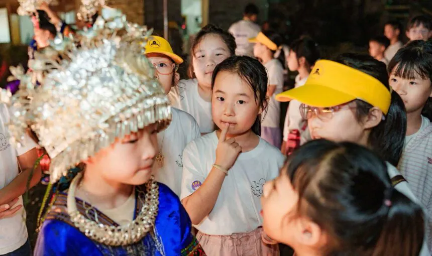 School trip in Guizhou, chinatrip, Children in a crowded festival, a girl in blue traditional dress with a beaded headdress among peers wearing yellow caps and white shirts.