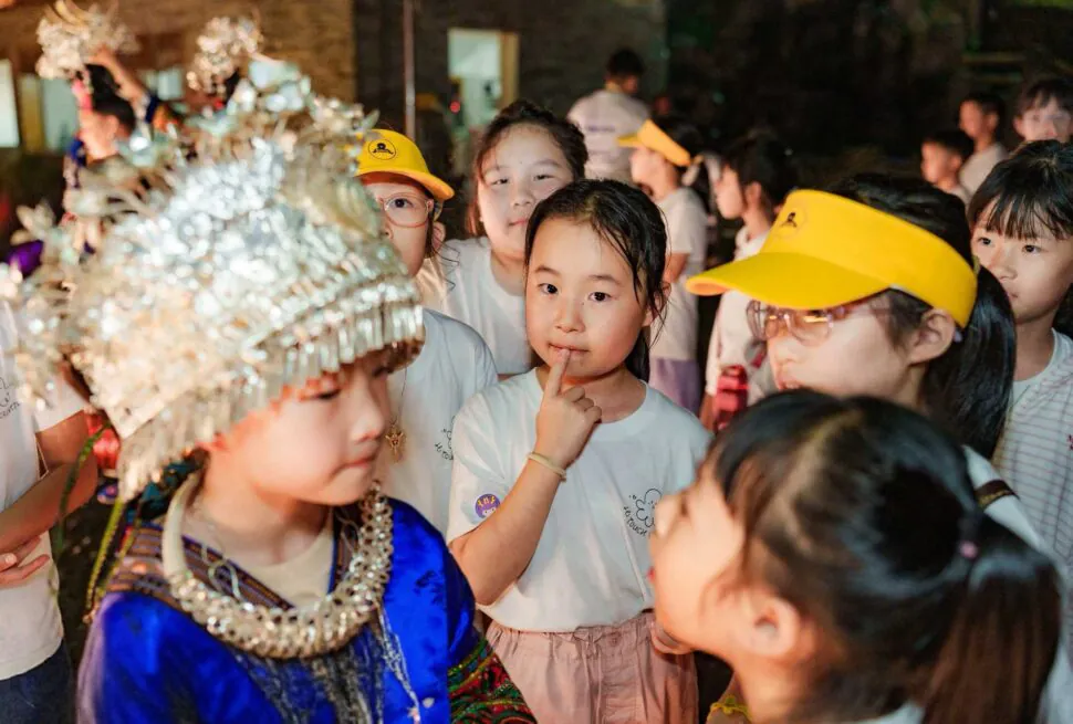 School trip in Guizhou, chinatrip, Children in a crowded festival, a girl in blue traditional dress with a beaded headdress among peers wearing yellow caps and white shirts.