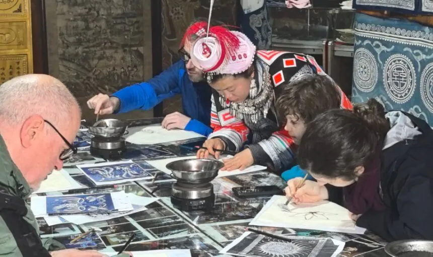 School trip in Guizhou,Chinatour Group of people around a table sketching on paper in a workshop, with blue patterned textiles in the background.