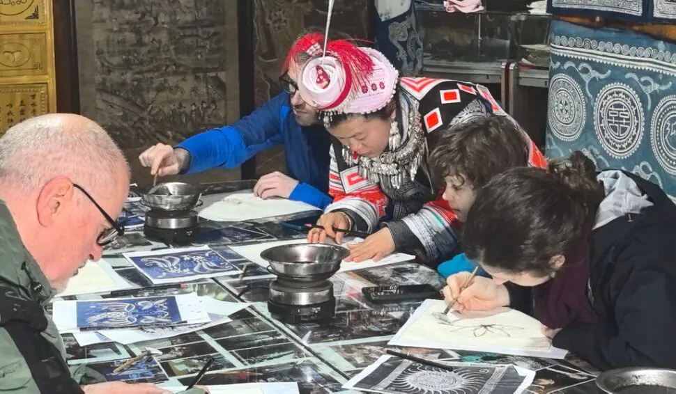 Group of people around a table sketching on paper in a workshop, with blue patterned textiles in the background.