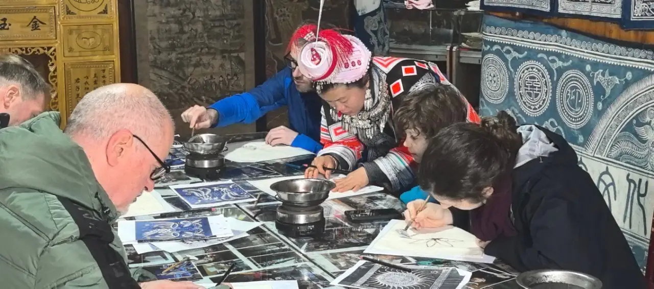 Group of people around a table sketching on paper in a workshop, with blue patterned textiles in the background.