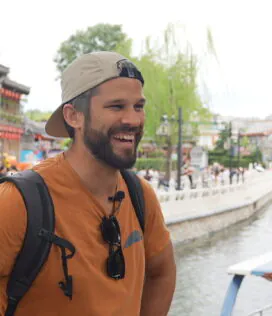China explorer tour, chinatravel, Smiling man with a backpack and sunglasses on his chest stands near a canal in a traditional Asian town, wearing a tan cap.