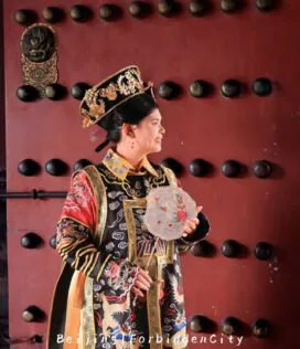 hanfu photoshoot, the forbidden city, beijing, qing dynasty, female, solo, chinatrip, Young woman in an ornate Chinese imperial costume with a jeweled headdress, holding a lace-edged fan against a red studded door.