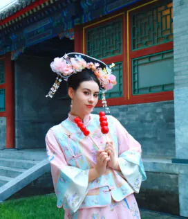 hanfu photoshoot, the forbidden city, beijing, qing dynasty, female, solo, chinatravel, Woman in pastel hanfu with a large flower headdress posing with red candy skewers in front of a traditional Chinese building.