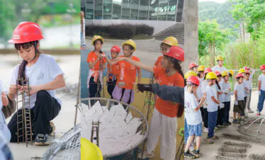 school trip china guizhou, chinatrip, Group of children in orange and white shirts wearing red and yellow hard hats collaboratively assembling a wire-frame sculpture outdoors.