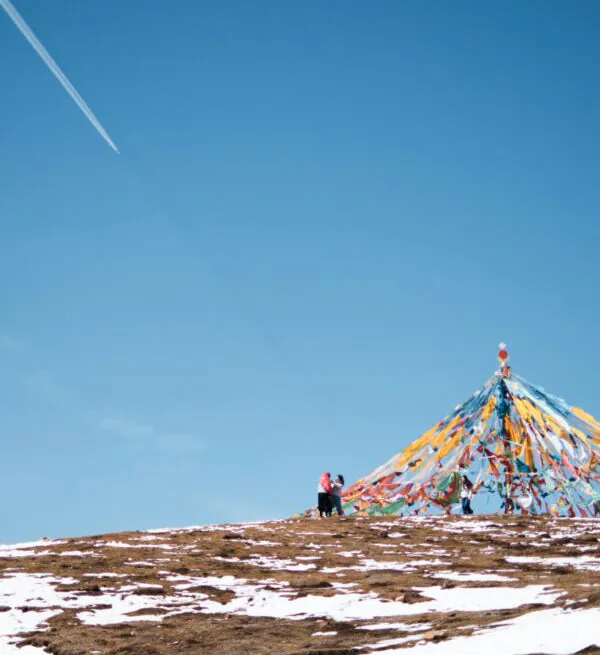 Dongda Mountain Pass,Tibet,Chinatour Two people stand near a colorful prayer flag pyramid on a snow-dusted hill against a clear blue sky.