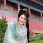 hanfu photoshoot, the forbidden city, beijing, female, solo, chinatravel, Smiling woman in pale blue traditional Hanfu holding a lace fan, posing outdoors near a temple/ Forbidden City backdrop.
