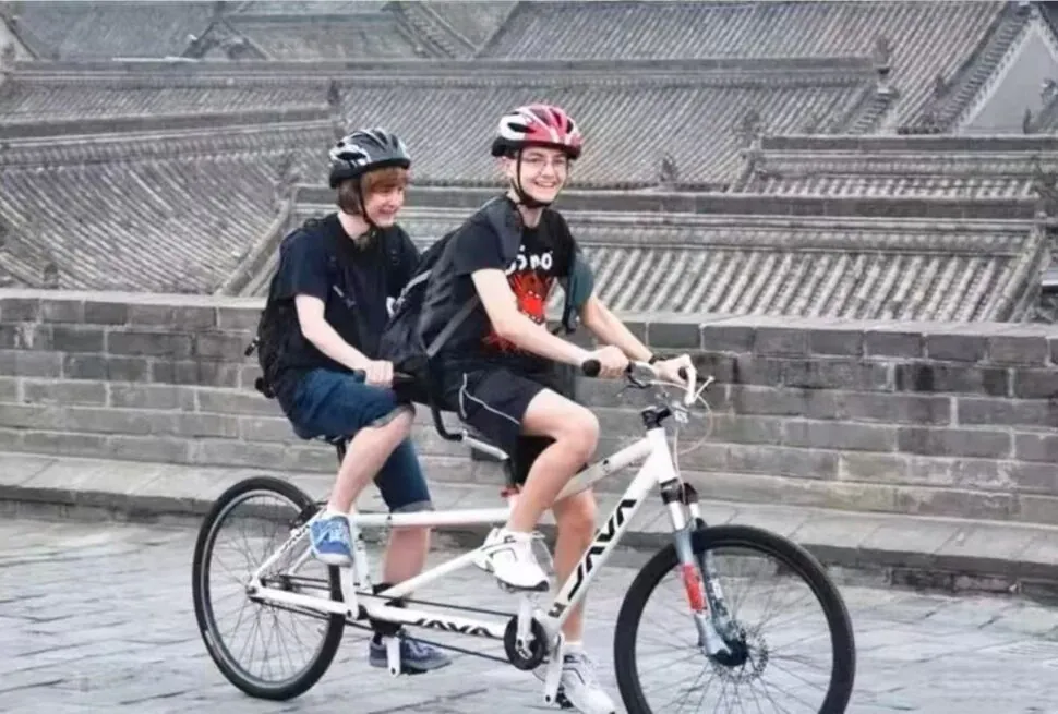 Two people ride a white tandem bicycle wearing helmets along a stone-paved street with a stone wall in the background (front rider smiling).