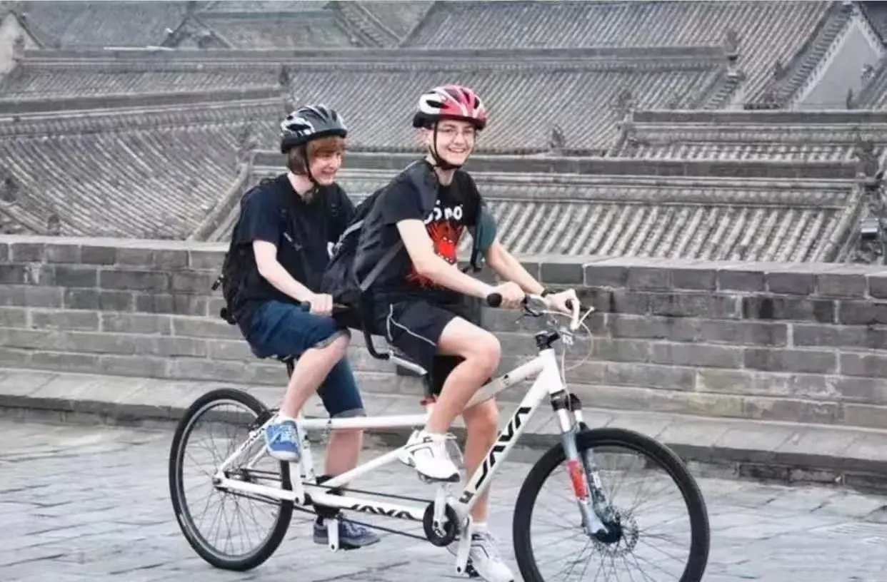 Two people ride a white tandem bicycle wearing helmets along a stone-paved street with a stone wall in the background (front rider smiling).