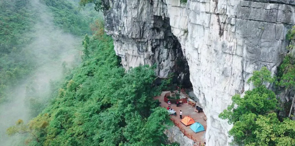 School trip in Guizhou, chinatrip, Cliffside campsite on a rocky ledge with colorful tents and seating, surrounded by misty green forest below a sheer rock face.