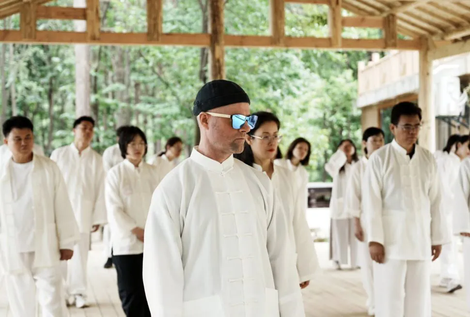 taichi wudang, hubei, chinatravel, Group of people in white traditional uniforms standing in an outdoor wooden pavilion, front man wearing a black cap and blue mirrored sunglasses, likely practicing tai chi.