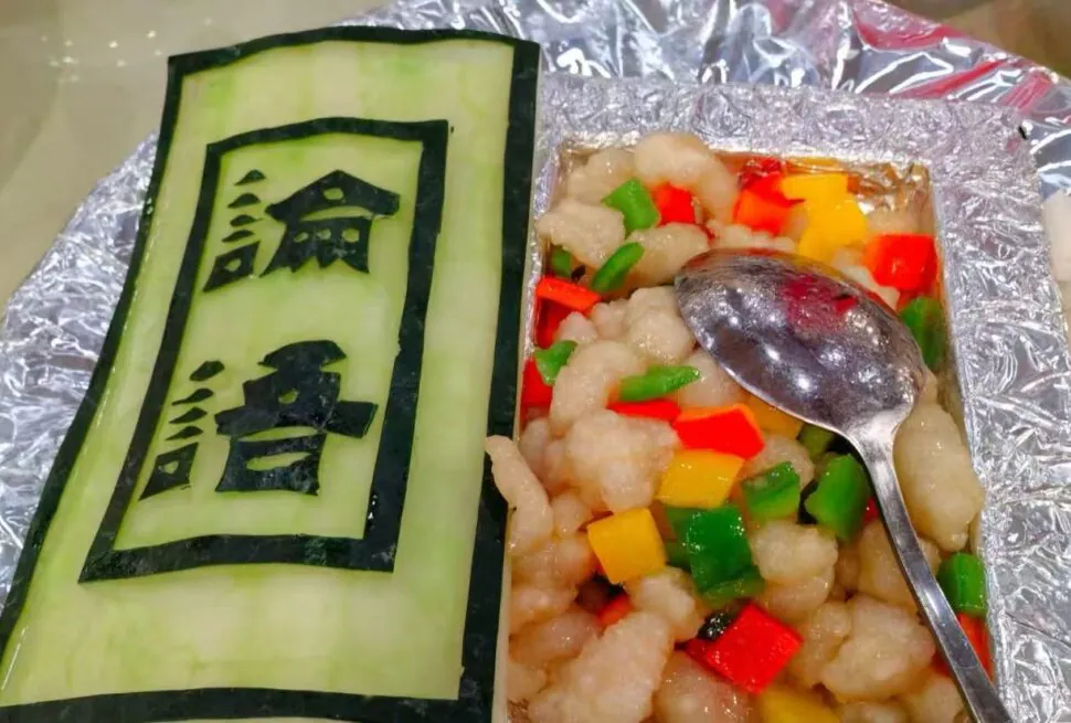 Green cucumber sheet with bold black Chinese characters beside a foil tray of diced red, yellow, and green peppers mixed with pale chicken, with a spoon resting on top.