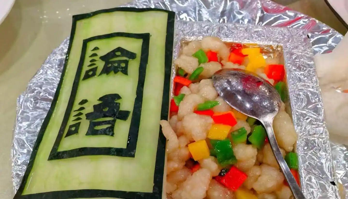 Green cucumber sheet with bold black Chinese characters beside a foil tray of diced red, yellow, and green peppers mixed with pale chicken, with a spoon resting on top.