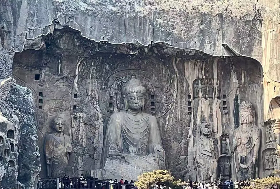 Large central Buddha statue carved into a rock cliff with smaller surrounding figures; visitors at the base for scale.