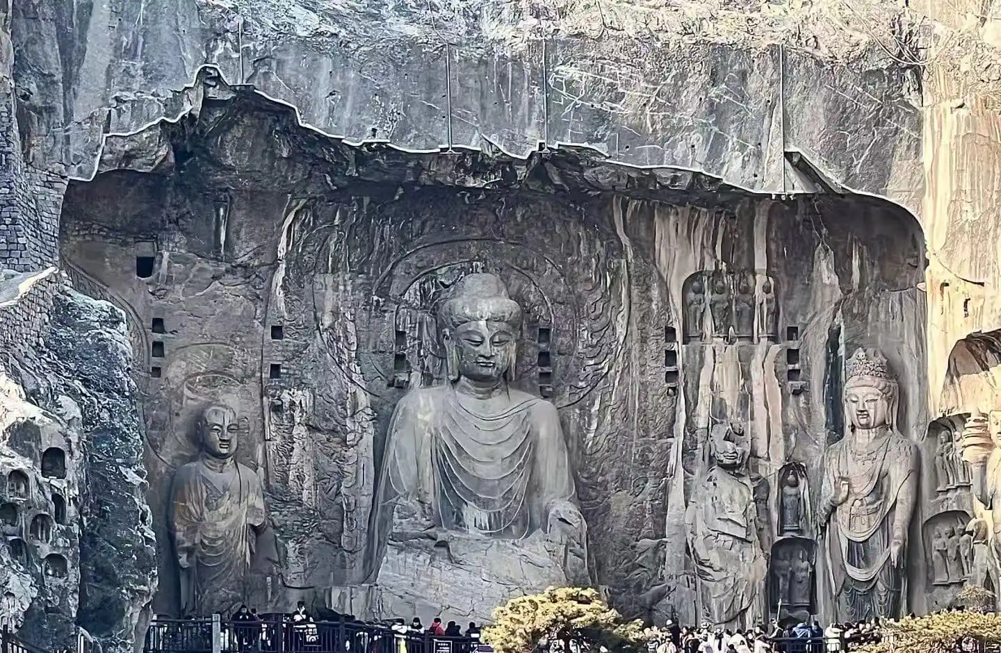 Large central Buddha statue carved into a rock cliff with smaller surrounding figures; visitors at the base for scale.
