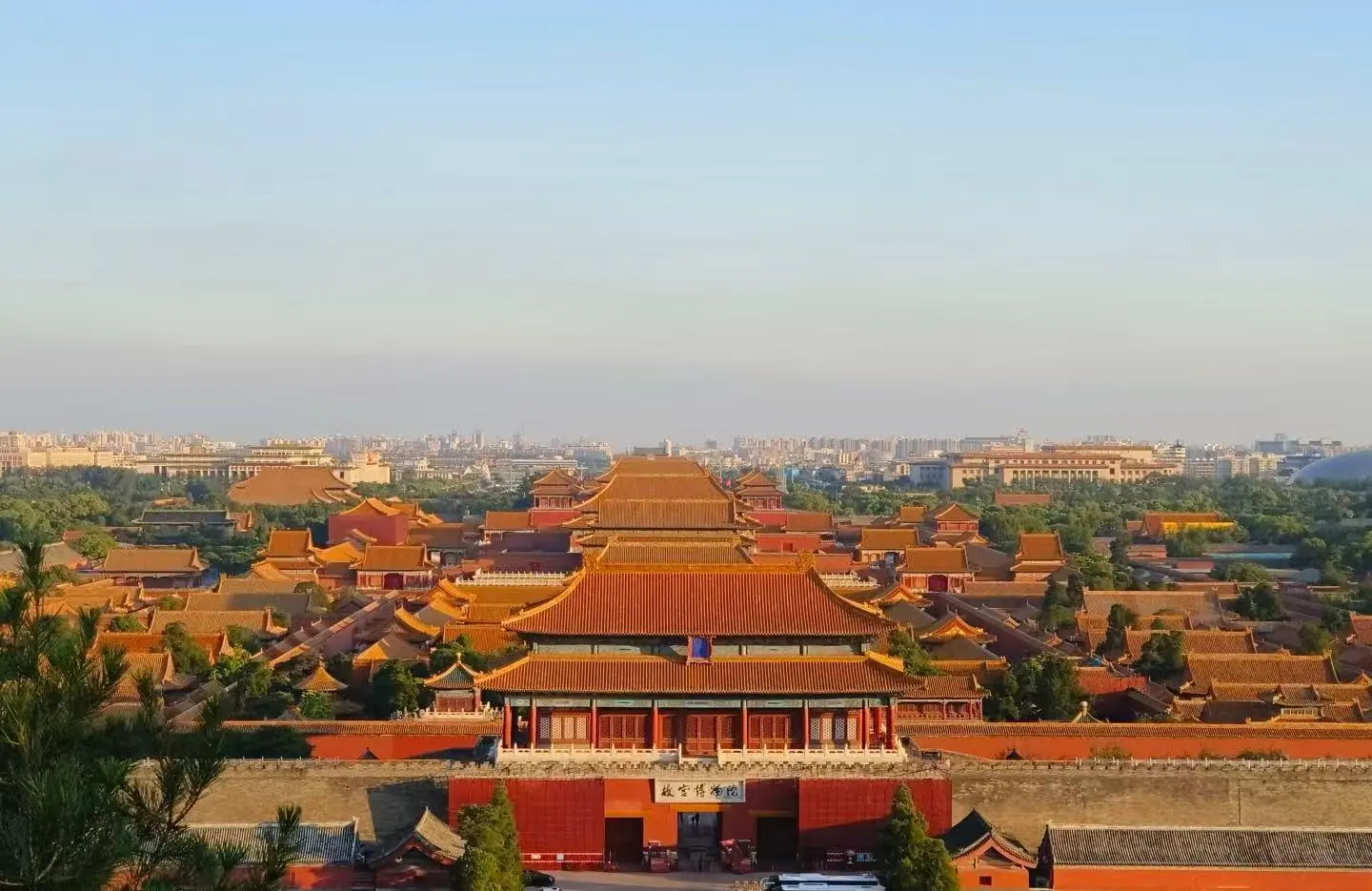 Panoramic view of the Forbidden City in Beijing, with red walls, golden-tiled roofs, and a distant city skyline on the horizon.