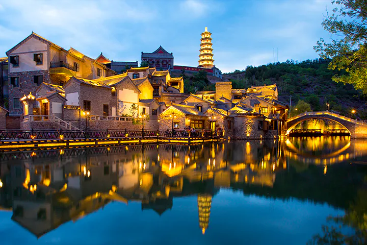 Evening view of a Chinese riverside town with lit traditional houses along a calm canal, a stone arched bridge, and a tall pagoda reflected in the water.