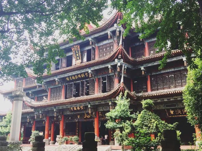 Multi-story Chinese temple with curved tiled roofs, red columns, gold plaques, and surrounding greenery.