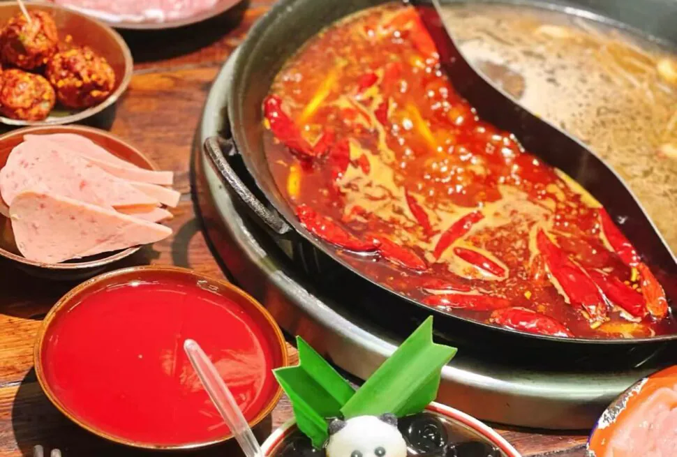 Hot pot with spicy red chili broth and peppers, surrounded by sliced meat, meatballs, and a red dipping sauce on a wooden table.