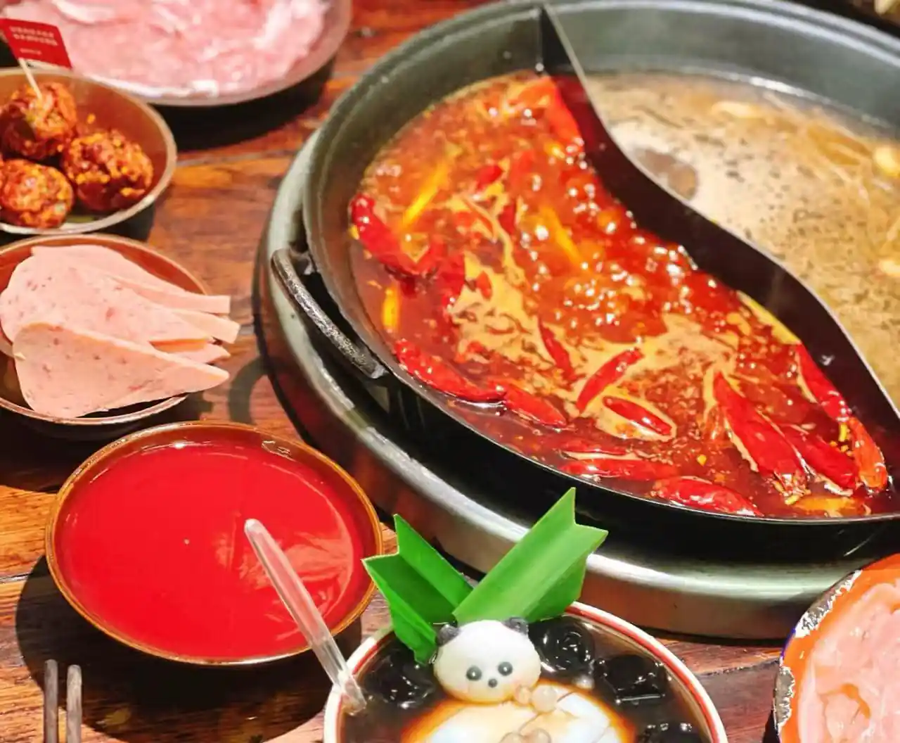 Hot pot with spicy red chili broth and peppers, surrounded by sliced meat, meatballs, and a red dipping sauce on a wooden table.